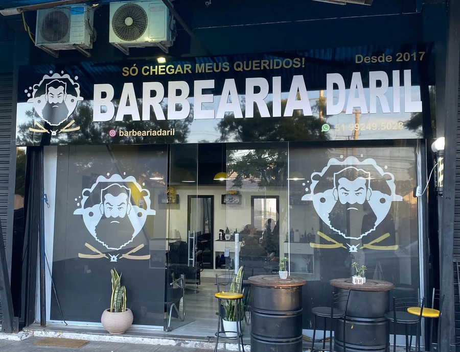 A fachada da Barbearia Daril de dia: toldo azul, letreiro preto e amarelo, tambores e banquetas na calçada.